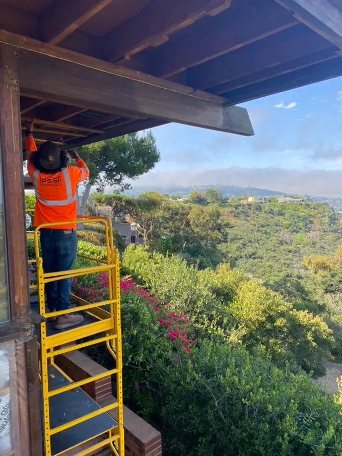 A worker on a yellow platform ladder works at the underside of the existing deep roof overhang, canyon brush and a distant ridge behind him.