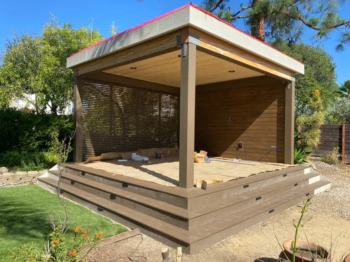 Seen from the lawn at the outside corner, where both stair runs meet and the louvered screen shows on the left face. The deck is still under protection paper, and the roof edge still shows underlayment above the fascia.