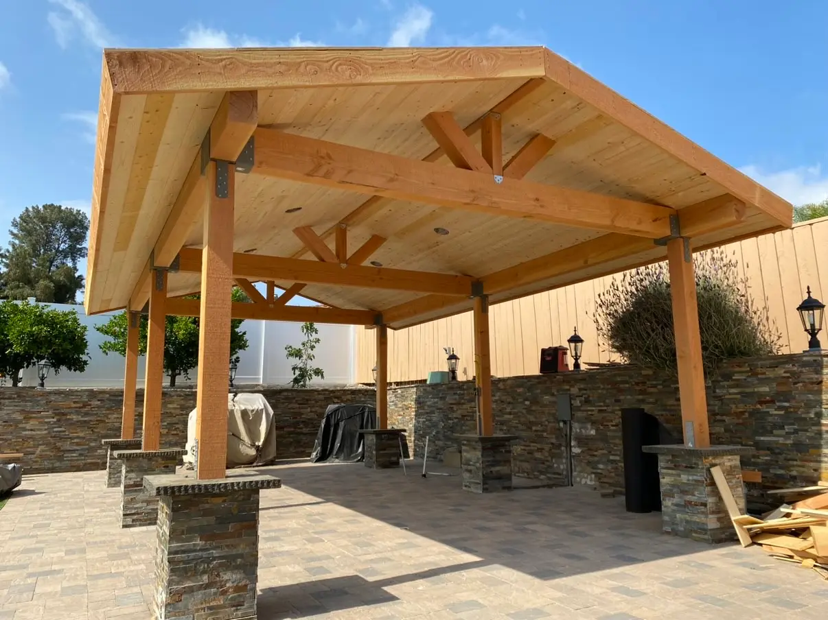 Open cedar pavilion with exposed trusses over a paver patio and outdoor kitchen