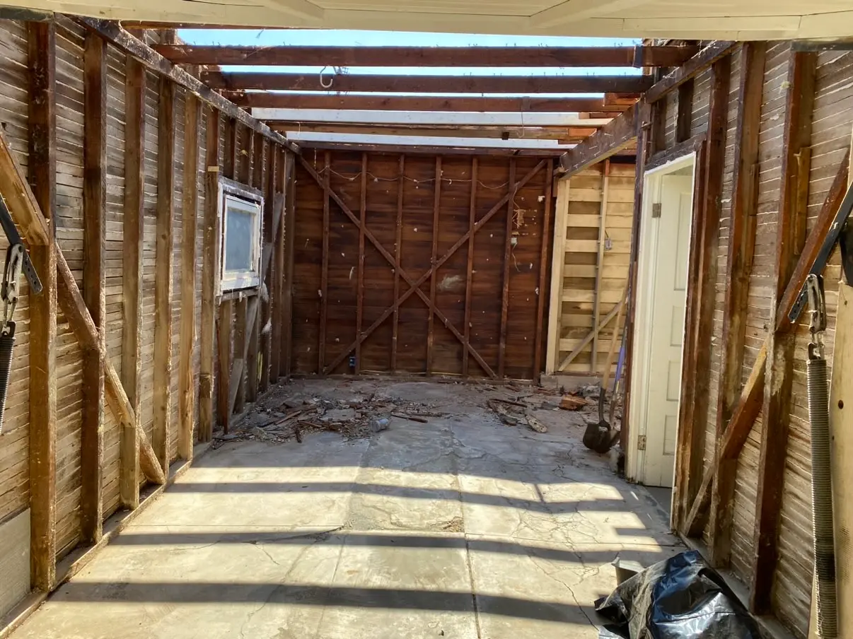 The garage stripped back to its original studs and let-in diagonal bracing. The roof decking is already off, so daylight comes through the rafters onto the bare slab.